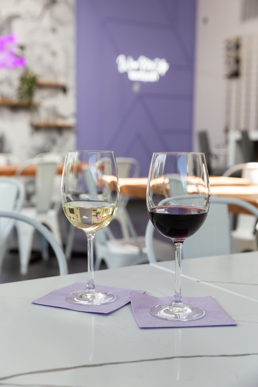Two wine glasses at Wine Me Up — one white, one red — on a table with the glowing neon sign behind them